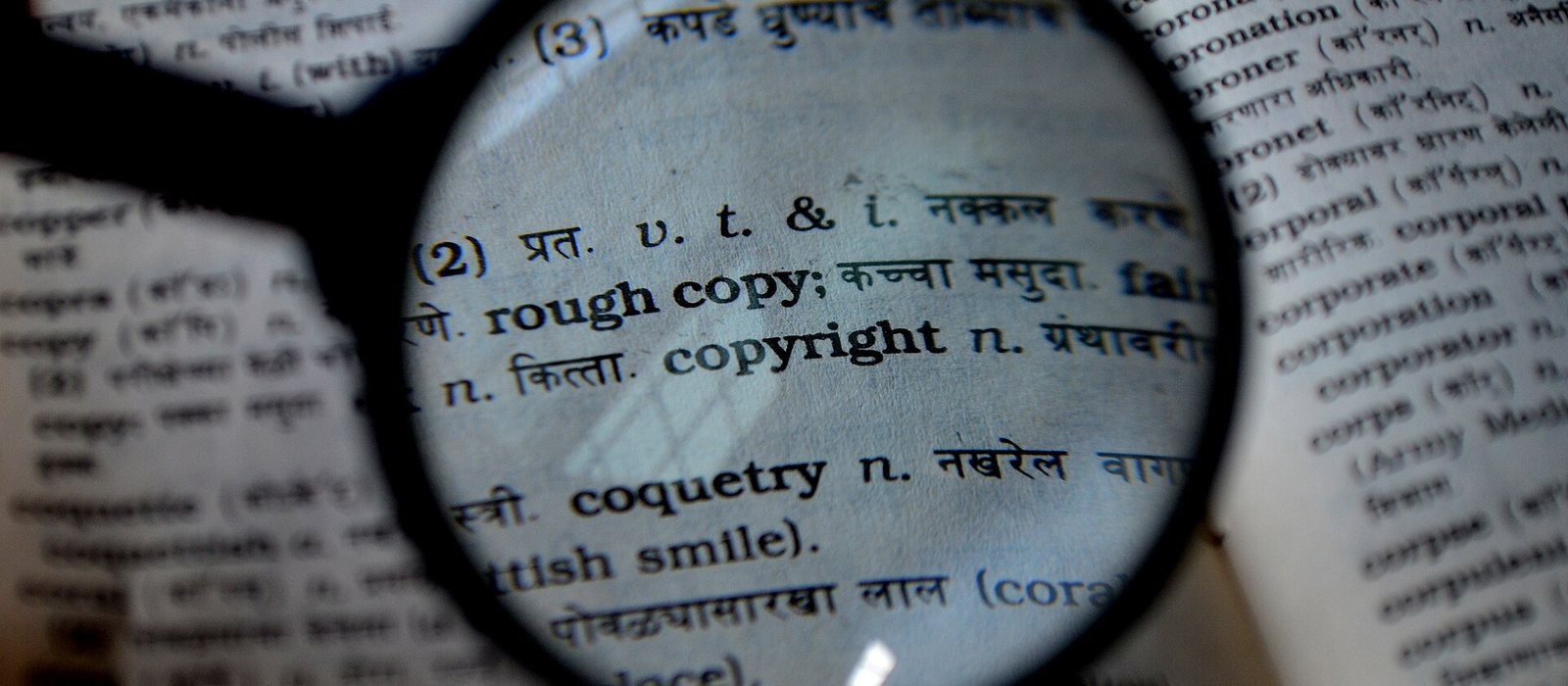 Lupe über einem aufgeschlagenen Wörterbuch, vergrößert sind die Einträge „rough copy“ und „copyright“